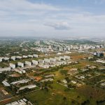 Vista aérea de Brasília, com áreas verdes, vias e edifícios urbanos sob luz do dia.