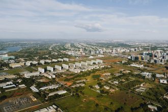 Vista aérea de Brasília, com áreas verdes, vias e edifícios urbanos sob luz do dia.