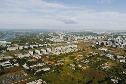 Vista aérea de Brasília, com áreas verdes, vias e edifícios urbanos sob luz do dia.
