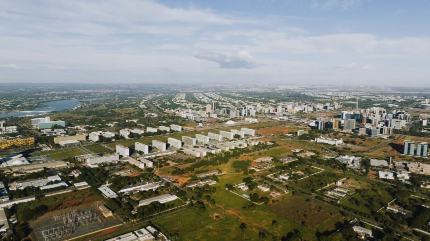Vista aérea de Brasília, com áreas verdes, vias e edifícios urbanos sob luz do dia.